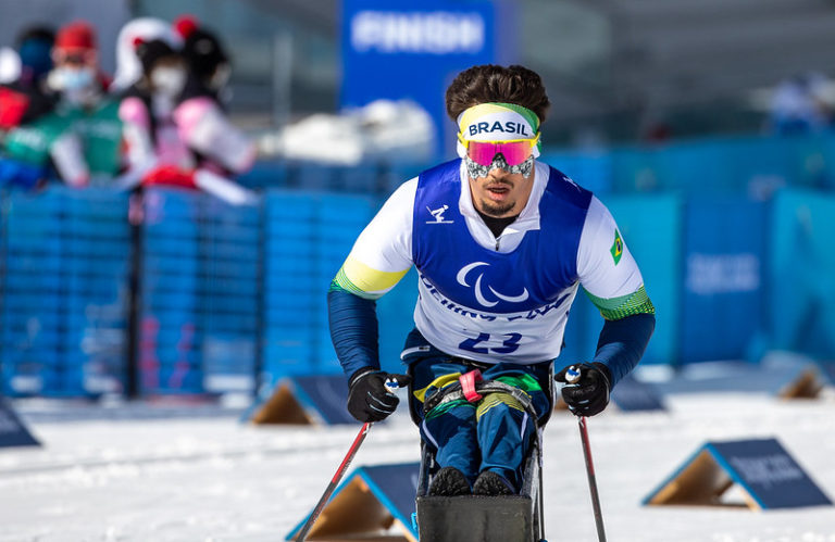 Homem com deficiência está esquiando na neve. Com óculos e roupas apropriadas para o frio, o atleta pratica esqui sentado.