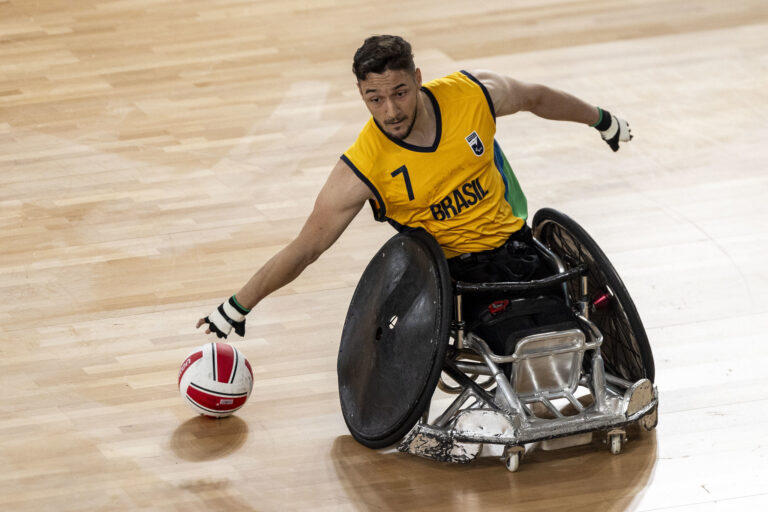 Um atleta do Brasil joga rúgbi em cadeira de rodas. Ele está esticando o braço para alcançar a bola que está no chão.