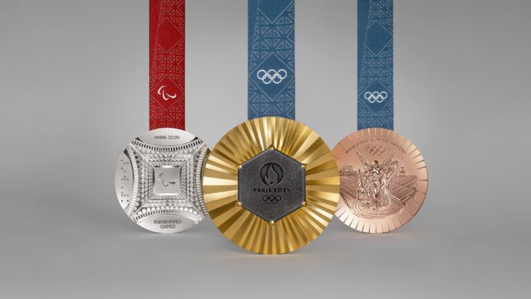 A imagem mostra três medalhas: uma de ouro, uma de prata e uma de bronze. Duas delas têm fita azul com os arcos olímpicas. Outra tem fita vermelha com estampa dos agitos paralímpicos. Elas serão distribuídas nos Jogos de Paris 2024
